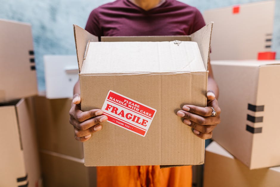 A person holding an open cardboard box with a red and white 'Fragile' handling label, standing inside a moving or storage environment. The box appears to be made of brown corrugated cardboard with taped seams and is empty at the top. The individual’s hands are visible, with one wearing a ring on the ring finger, and their body is partially obscured behind the box. In the background, there are several other cardboard boxes, some sealed with black packing tape, stacked on a surface or against a wall, indicating an organized packing or furniture transport setting typical in home relocation or house removals. The scene is lit with neutral lighting, emphasizing the focus on safe packing practices and careful handling of delicate items, aligning with professional moving services such as those provided by Haringey Removals.