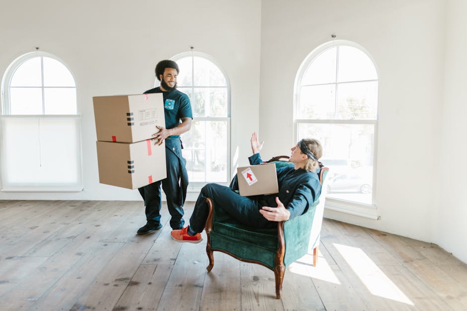 A professional moving team inside a bright, spacious room with white walls and large arched windows, where one mover is carrying two cardboard boxes secured with red and black tape, smiling and positioned near the window, while a resident or client, dressed casually, is seated in an upholstered armchair with wooden legs. The seated person is holding a smaller box with a red arrow sticker on it, raising one hand in a greeting or dismissal gesture, and appears to be engaging with the mover. The room has wooden flooring and minimal furnishings, indicating a home relocation or packing process. The scene captures the loading or unpacking stage of a house move, with the movers and resident working together during a furniture transport or moving service, supported by Haringey Removals as part of their removals service category.
