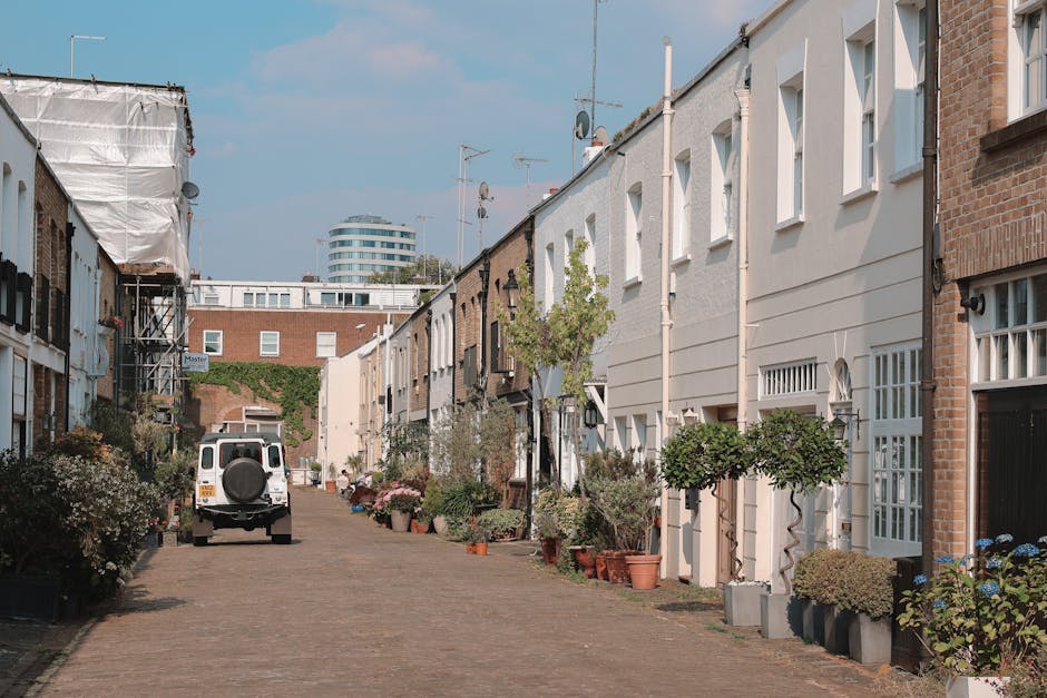 A narrow residential street during daytime with brick and white-painted terraced houses on both sides, each with small front gardens filled with potted plants and trees. The street has a cobblestone surface with a white van parked near the middle, positioned to assist in a home relocation or furniture transport process. Some houses display exterior features such as black lantern-style lighting fixtures and window shutters. There is visible scaffolding on the left side of the street, indicating ongoing building work. The sky is partly cloudy with a few clouds, and in the background, a modern multi-storey building can be seen behind the row of houses. The scene depicts an environment suitable for a professional removals company, such as Haringey Removals, handling packing, loading, and transportation activities in tight urban settings typical of Crouch End and similar areas.