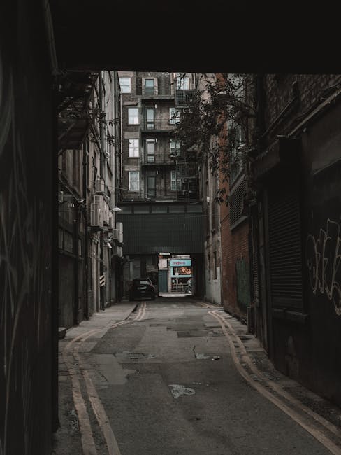 A narrow urban alleyway in Crouch End, Haringey, showing a paved path bordered by dark brick and metal-clad buildings on both sides. On the left, there are scaffolding and a metal staircase attached to the building, with a few small air conditioning units and graffiti visible on the walls. On the right, a closed roller shutter door with graffiti is present, and a small window nearby. The alley is dimly lit with limited natural light, leading towards a parked car at the end, which is positioned near a recessed loading area with a loading bay sign above. In the background, a taller residential building with multiple balconies and windows is visible, suggesting a dense city environment. This setting reflects typical challenges faced during home relocation in tight city streets, where careful planning for furniture transport, loading, and parking is essential. Haringey Removals offers professional moving services suitable for such narrow street and parking constraints, supporting smooth packing and loading processes during house removals.