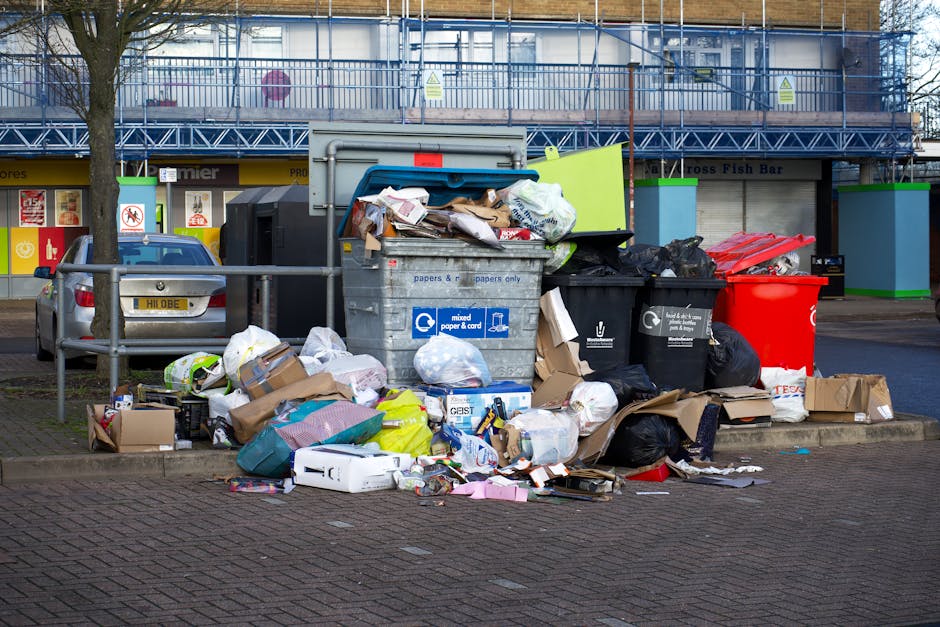 A collection of overflowing rubbish and recycling bins situated on a paved sidewalk area outside a commercial building in Haringey. The scene includes a large grey mixed paper and card recycling bin with its lid partially open, revealing flattened cardboard boxes, paper, and packaging materials. Surrounding it are several black, green, and red waste bins, some filled with plastic bags and disposed items. Cardboard boxes and extra garbage are scattered on the ground, with some items leaning against the bins. A silver car is parked nearby, and a metal railing separates the waste area from the street. In the background, a building under renovation features scaffolding, a yellow storefront, and signage, indicating typical urban environment elements related to property and house removals, emphasizing the importance of proper waste disposal during home relocation or furniture transport processes.