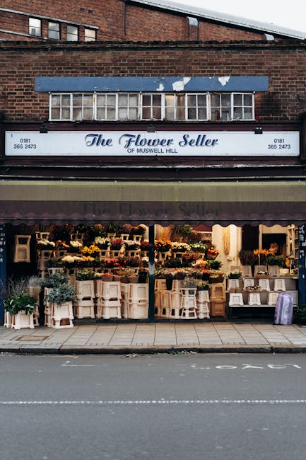 The image shows the storefront of 'The Flower Seller of Muswell Hill,' a florist shop on an urban street with a brick building above. The shop has a navy awning with the business name printed in white and purple, partially covering the display area. Outside, various potted plants, flowers in buckets, and floral arrangements are arranged on white wooden display tables and shelves, creating a vibrant and colourful scene. The sidewalk in front is clean and extends along the shop frontage, with a visible curb separating it from the road, which features lane markings. Inside the shop, visible through large glass windows, are additional floral supplies and arrangements. The setting appears to be during evening or late afternoon, as the interior lights cast a warm glow. This image is relevant to house removals and relocation services, illustrating the type of local businesses that may require moving or logistical support during a home relocation process, as provided by companies like Haringey Removals.