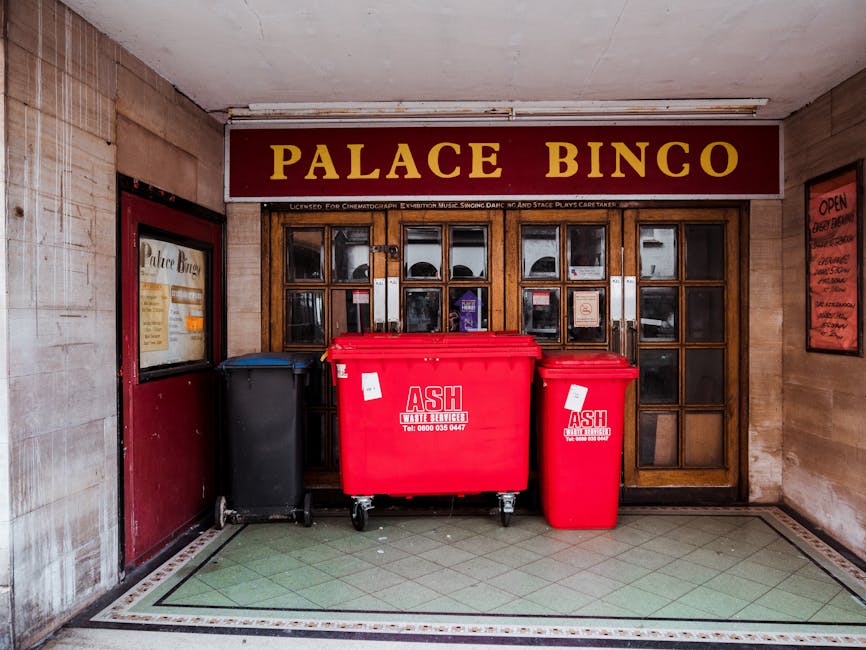 A collection of overflowing rubbish and recycling bins situated on a paved sidewalk area outside a commercial building in Haringey. The scene includes a large grey mixed paper and card recycling bin with its lid partially open, revealing flattened cardboard boxes, paper, and packaging materials. Surrounding it are several black, green, and red waste bins, some filled with plastic bags and disposed items. Cardboard boxes and extra garbage are scattered on the ground, with some items leaning against the bins. A silver car is parked nearby, and a metal railing separates the waste area from the street. In the background, a building under renovation features scaffolding, a yellow storefront, and signage, indicating typical urban environment elements related to property and house removals, emphasizing the importance of proper waste disposal during home relocation or furniture transport processes.