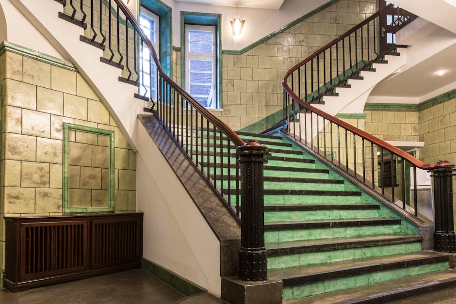 Interior view of a historic staircase within a residential building featuring green tiled steps and dark wooden handrails supported by black iron balusters. The staircase curves upward and is illuminated by natural light coming from multiple windows at the top, with an additional wall-mounted light fixture on the wall above. The walls are adorned with beige ceramic tiles arranged in a rectangular pattern, framed by green trim at the top and bottom, and there is a wooden radiator cover positioned against the lower part of the wall. The setting appears to be prepared for a home relocation or furniture transport process, with visible space for moving boxes or furniture pieces. The scene provides insight into the environment where professional removals, like those offered by Haringey Removals, might operate during stair access and loading procedures for house removals in the N22 Wood Green area.