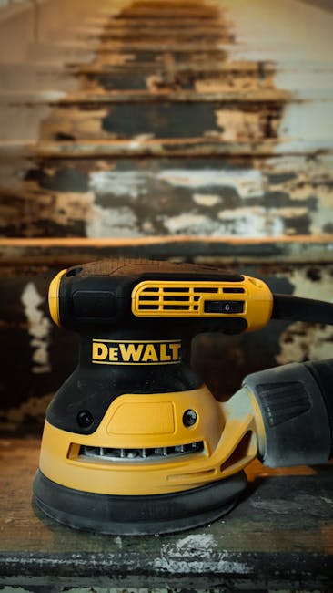Close-up of a yellow and black DeWalt electric sander resting on a wooden staircase landing, with the stairs above showing significant peeling paint and worn wood surfaces. The sander is positioned at the bottom of the image, with its cord extending out of view, indicating it is being used for sanding or preparing the staircase for painting or refurbishment. The staircase is partially visible, with the worn steps revealing layers of old paint and wood, suggesting ongoing renovation or home improvement work as part of a house removal or refurbishment project. The environment appears indoors, with natural or overhead lighting highlighting the textured, aged staircase surface. This scene illustrates the preparation process involved in property relocation or renovation managed by companies like Haringey Removals, supporting services including packing, furniture transport, and home refurbishment.
