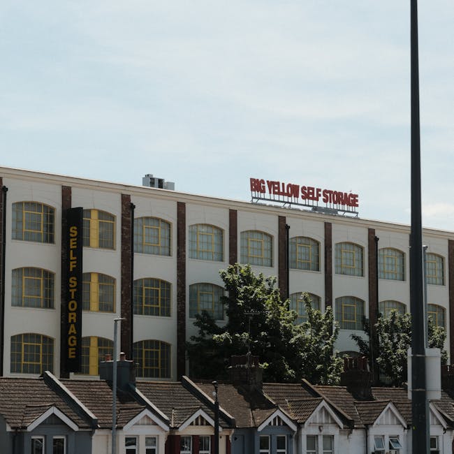 A multi-storey building with a white facade and large, arched yellow-framed windows hosting a self-storage facility. The sign on the building reads 'Big Yellow Self Storage' in red letters on the roof and 'Self Storage' vertically on a black strip on the side. Below the building, there are small, row houses with sloped tiled roofs and chimneys. Trees partially obscure the view of the lower part of the building, and a streetlamp is visible on the right side of the image. The scene appears to be taken during daytime with clear skies, indicating an urban environment potentially involved in house removals or furniture transport activities, as relevant to services offered by Haringey Removals.