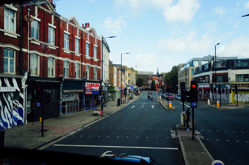 A street scene in Haringey showing a wide road with a loading area and pedestrian crossings. On the left side, there are brick buildings with commercial storefronts, including a shop with graffiti-style artwork on its shutters and a sign for 'Carolina.' On the right side, modern white and grey apartment buildings with balconies and graffiti on walls are visible. The road has traffic lights and vehicles including a white van parked near the curb, suggesting an ongoing home relocation or furniture transport operation. Several pedestrians are waiting at the pedestrian crossings, and the scene is lit by natural daylight with a partly cloudy sky overhead. This setting illustrates typical urban moving logistics, with items such as boxes and furniture possibly being loaded or unloaded in the context of house removals facilitated by Haringey Removals.