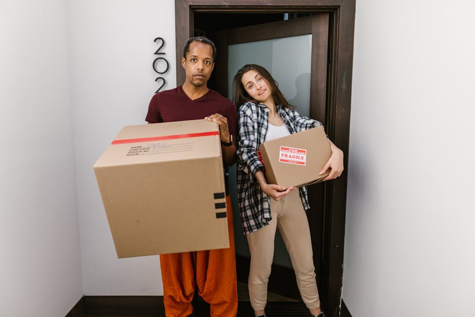 A man and a woman standing inside a narrow apartment entryway during a home relocation, each holding a cardboard box. The man, dressed in an orange pair of trousers and a maroon t-shirt, holds a large box with black tape and labels. The woman, wearing beige trousers and a black-and-white checkered shirt over a white top, holds a smaller, marked fragile box. Both are positioned near an open doorway with a dark wooden frame and frosted glass panel, leading into an interior space with white walls. The scene is lit by natural or indoor lighting, capturing the loading process involved in packing and moving, as handled by a professional removals company such as Haringey Removals, which specialises in furniture transport and moving logistics. The focus is on the careful handling of packed boxes, highlighting the importance of appropriate packing materials like cardboard and proper positioning during a domestic move.