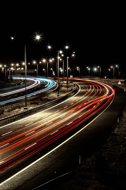 A long-exposure photograph of a busy road at night showing light trails created by vehicle headlights and taillights, with blue and white streaks from moving cars in the left lanes and red streaks from vehicles in the right lanes. The road curves to the right, and there are tall streetlights evenly spaced along the highway, illuminating the scene with bright white light. The surrounding area appears dark, with no visible buildings or pedestrians. This image highlights the flow of traffic during night-time home relocations and highlights the importance of carefully planning vehicle arrivals and departures, as might be relevant for professional removals like Haringey Removals, operating in areas such as Turnpike Lane.