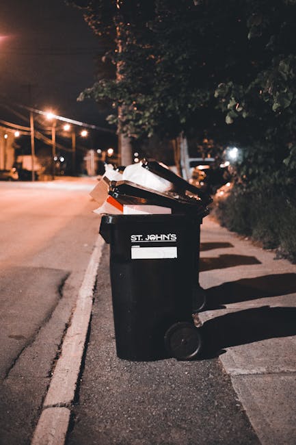 A row of waste bins with different coloured lids—red, yellow, and blue—lined up along the edge of a pavement next to a road. The blue bin, designated for recycling, is partially visible and appears to